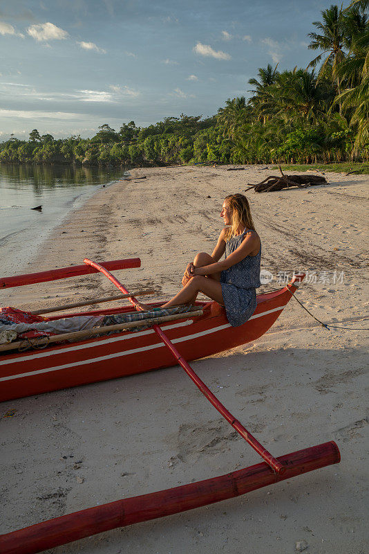 年轻女子坐在渔船上凝视夕阳，热带气候