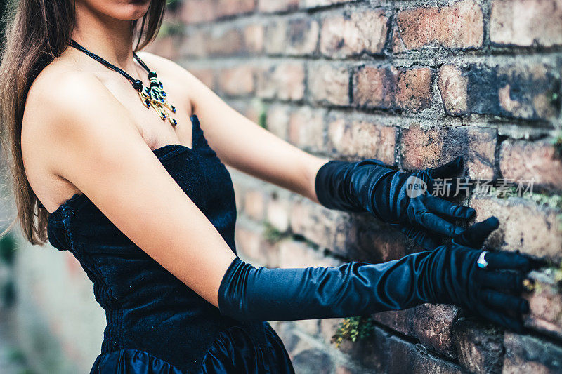 一个穿着黑裙子戴着手套的漂亮女孩站在一堵砖墙旁边