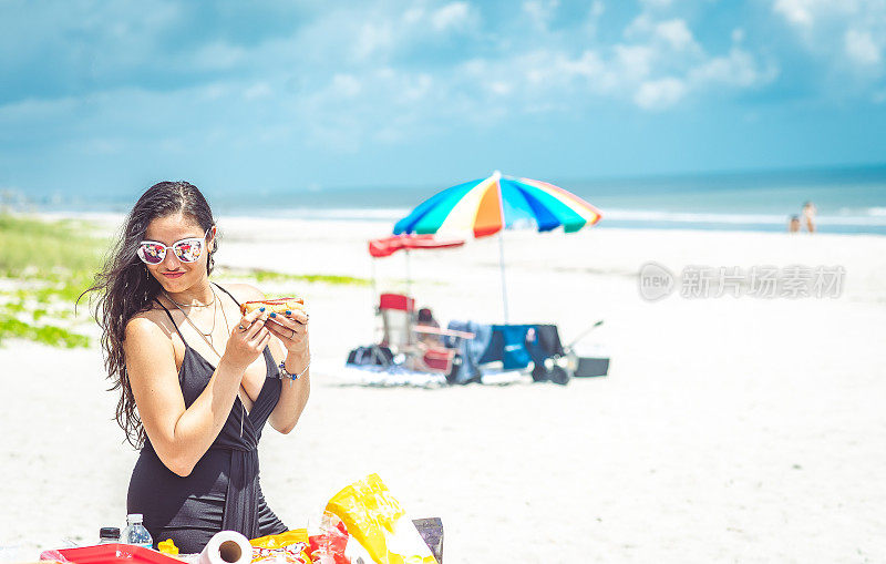 年轻漂亮的拉丁女人在海滩烧烤往热狗上涂番茄酱