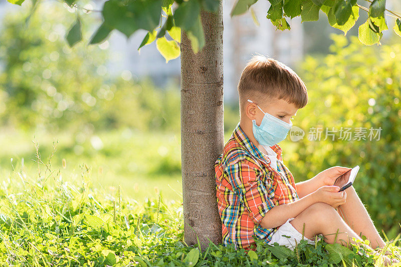 一个戴着医用口罩的孩子坐在草地上，看着夏日日落时的手机卡通。手里拿着手机的孩子。大流行期间的防疫工作