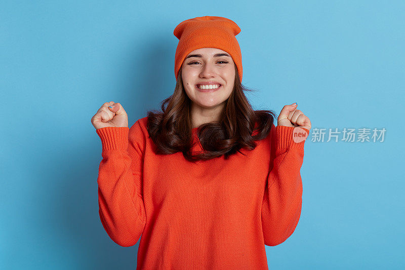 喜出望外的深色头发的欧洲年轻女士紧握两个拳头，庆祝成功和欢呼，做出欢呼的手势，胜利的好消息，穿着橙色毛衣和帽子。