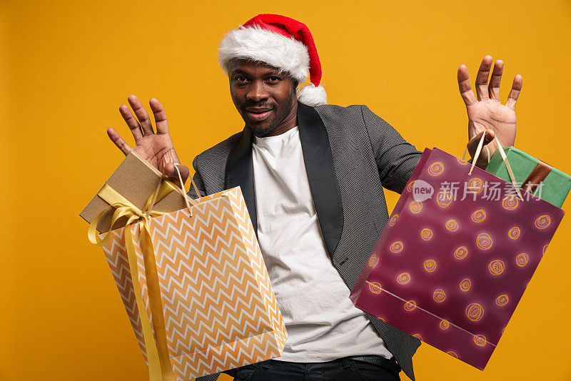 一个戴着圣诞帽、皮肤黝黑的男人举起双手，手里拿着装有礼物盒子的彩色袋子