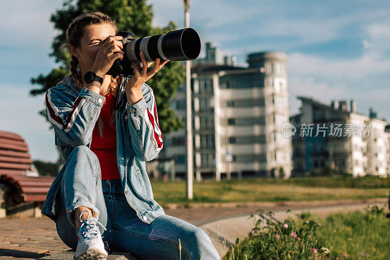 一位年轻的女摄影师正在户外的街道上工作。女孩坐在草地上拍照