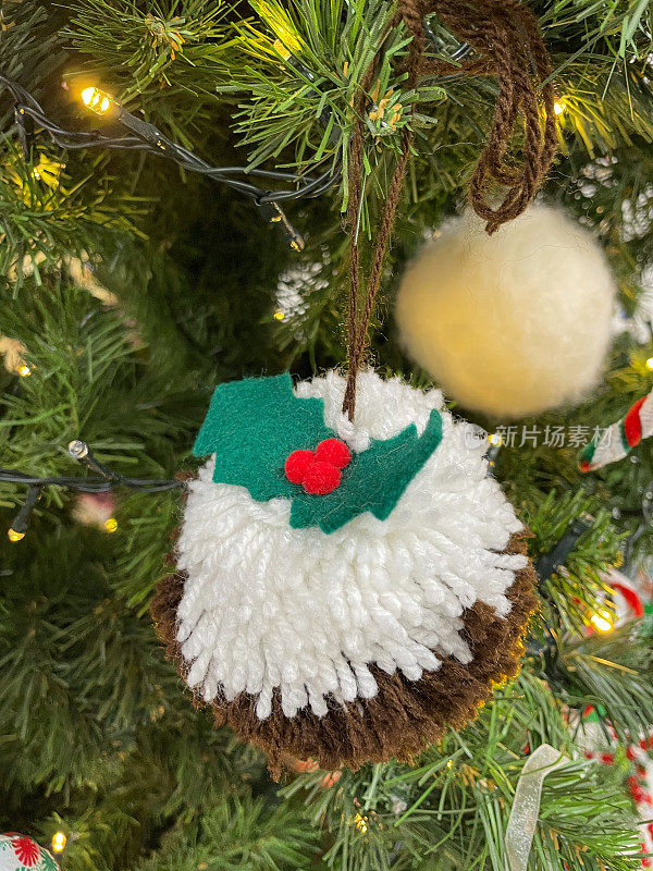 手工制作的羊毛圣诞布丁绒球在人造圣诞树树枝上的图像，绿色云杉针，模糊的白色精灵灯的背景