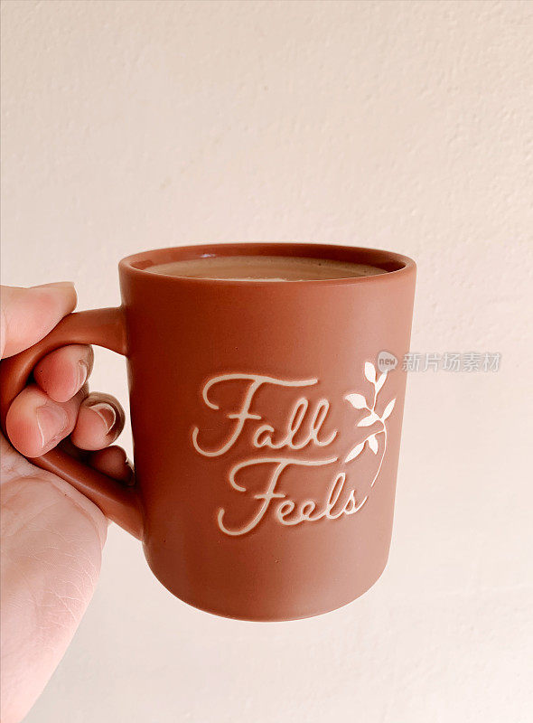 2022年的秋天，一名女子端着一杯热咖啡，杯子里有一个温暖的彩色陶土杯，上面写着“秋天的感觉”