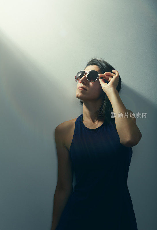 一个年轻的女人站在一个忧郁的环境的墙背景前，拿着她的太阳镜，看着光
