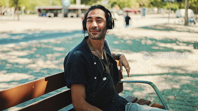 特写，年轻的意大利男子与马尾和胡茬坐在街头长凳上戴着耳机，听音乐在城市景观背景