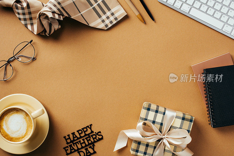 父亲节快乐的概念。用礼盒、键盘、领带、咖啡杯、纸质笔记本组成的平面构图，棕色背景上写着“父亲节快乐”
