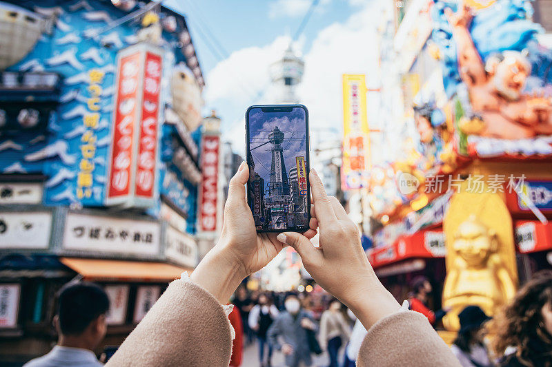 一位亚洲妇女正在日本大阪的津天阁观光。她用智能手机拍摄了津天阁岛和周围风景的照片。