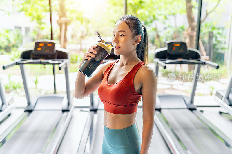 亚洲妇女休息，啜饮乳清蛋白在灼热的阳光下，在室内健身俱乐部与花园景观