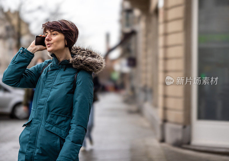 年轻女子在蓝色的雨衣在室外谈论她的手机在城市街道的人行道上