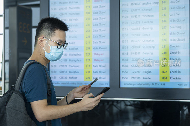 一名亚洲男子在机场的航班信息屏幕前，戴着医用外科口罩和背包，用手机和机票查看登机信息