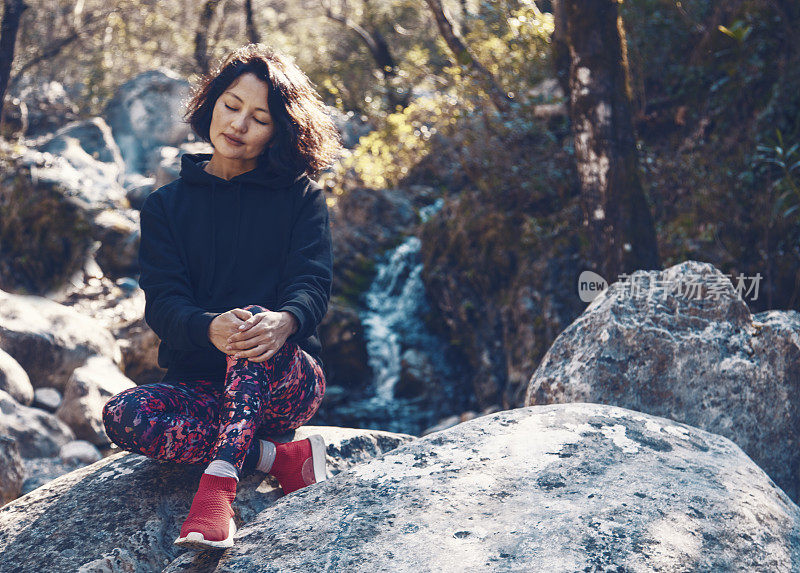 一名亚洲年轻女子，坐在一块岩石上，听着身后水流的声音，寻求平静。