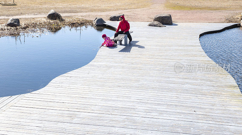 一位妇女带着孩子在湖边的公园里散步。一个女人坐在公园的长椅上，一个小孩用手摸着水。
