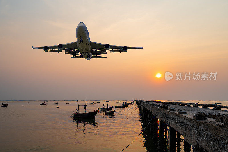 前方图像商用客机或货机飞越渔船，渔船在码头的海面上漂浮着，金色的日落海景