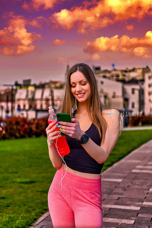一个年轻漂亮的女人站在城市的街道上，在五彩缤纷的天空下用手机。