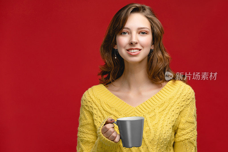 画室里的美女正拿着杯子喝饮料。概念上，人以红色为背景，人穿黄色套头衫，面带微笑。拿着咖啡或茶的女孩