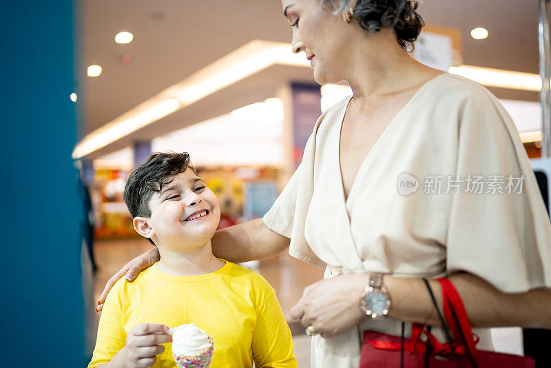 小男孩在吃冰淇淋，而他的妈妈在商场里看着橱窗