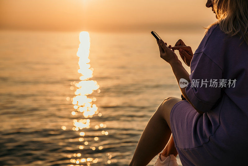 一名成年女子正被她的智能手机分心，而美丽的日落在海上，特写镜头