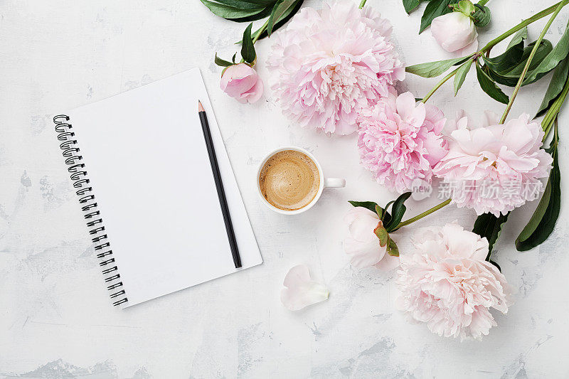 早餐用的咖啡杯，空笔记本，铅笔和粉红色的牡丹花。平的。女人工作的办公桌。