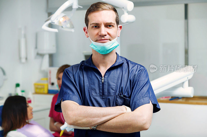 在牙科诊所里，年轻英俊的牙医双臂交叉站在镜头前。