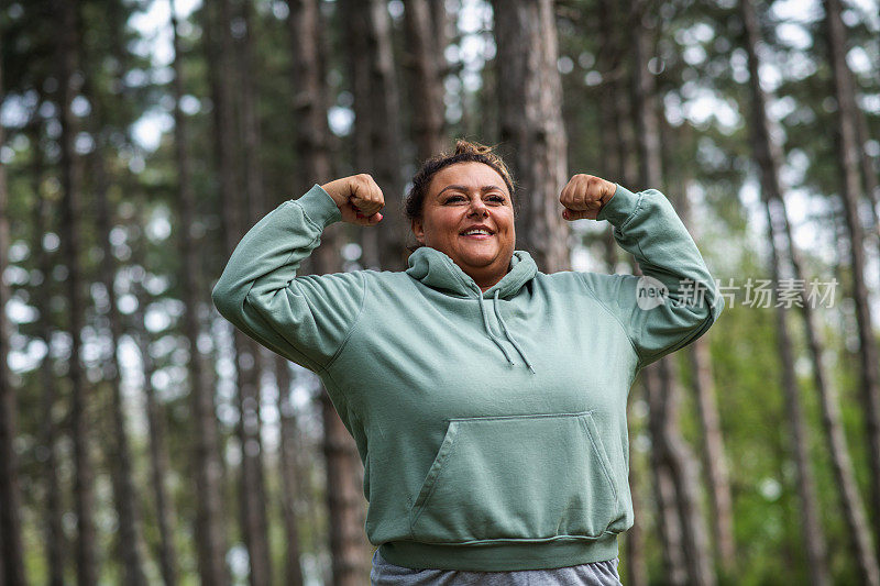 微笑的肥胖西班牙拉丁妇女在森林中弯曲手臂肌肉