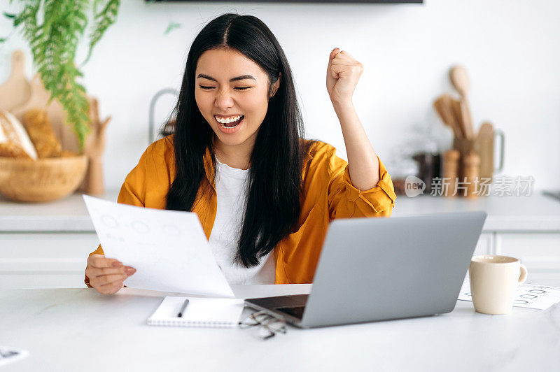 兴奋惊讶快乐的亚洲成功女性，一个公司员工，一个自由职业者，在笔记本电脑工作，手里拿着一个文件，高兴于大利润，在工作上的晋升，签订了一份利润丰厚的合同