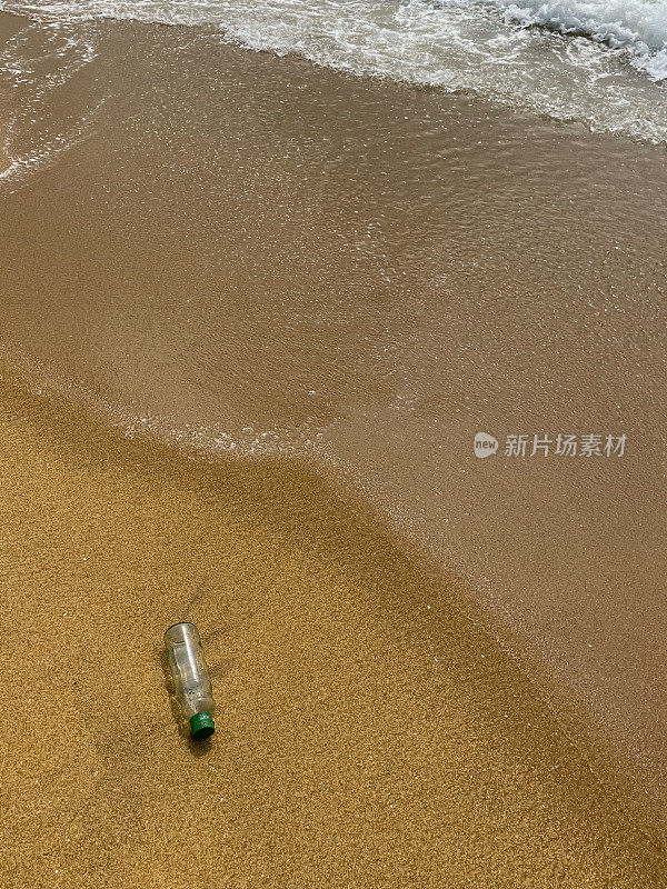 图片:空的，透明的塑料饮料瓶，绿色的盖子被冲到退潮水边缘的沙滩上，海浪把海洋垃圾和污染冲到岸上，乱扔的沙子，肮脏的海滩，高架视图，复制空间