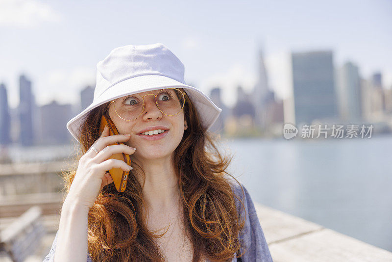 一名年轻女子在纽约猎人点的Gantry广场州立公园的码头上打电话，身后是曼哈顿的景色。