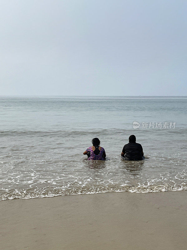 印度男子和女子在果阿海滩度假的照片，坐在海里，印度果阿帕洛莱姆，度假游客在海滩上洗澡，退潮时，后视图，重点在前景