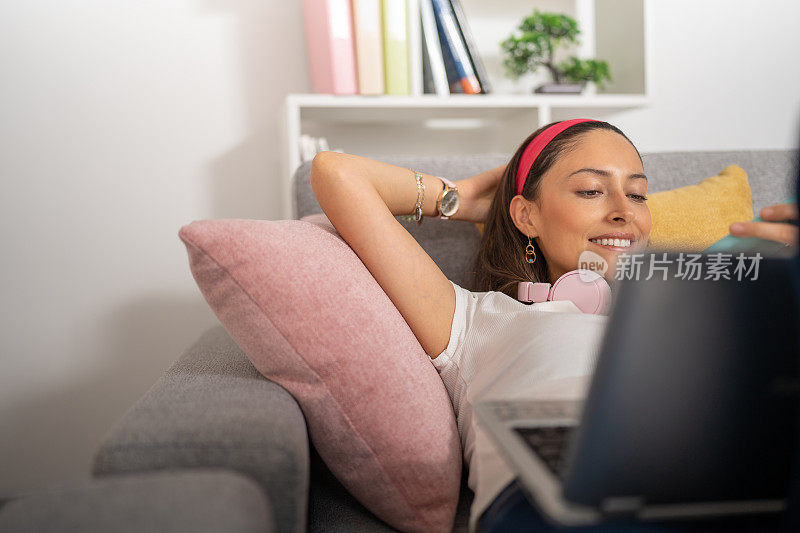 年轻女子，躺在沙发上讲手机，脖子上戴着耳机，头上放着笔记本电脑。