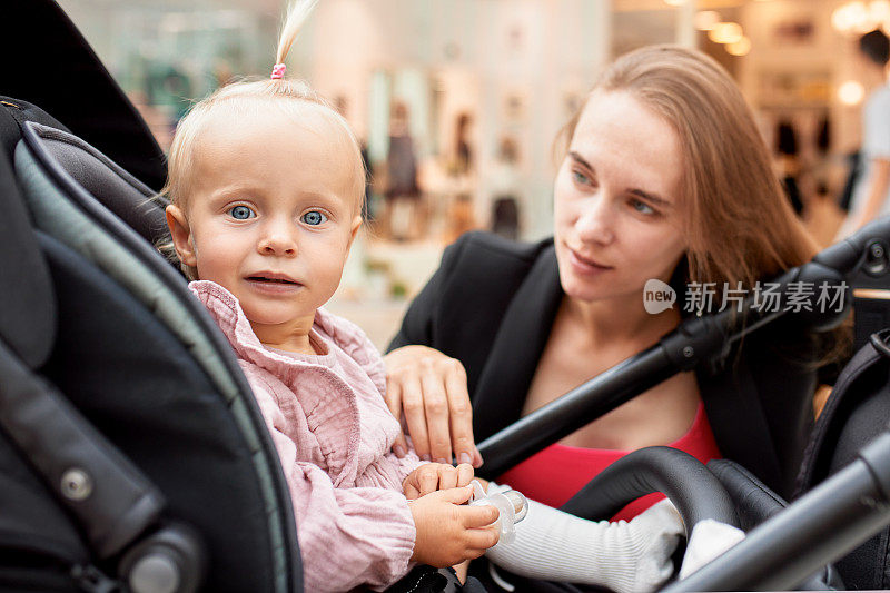 一个坐在马车里的小女孩的特写，金发碧眼的小女孩穿着粉红色的衣服。一位推着婴儿车的妈妈在商场里走来走去，坐在婴儿车旁边和她的女儿说话。