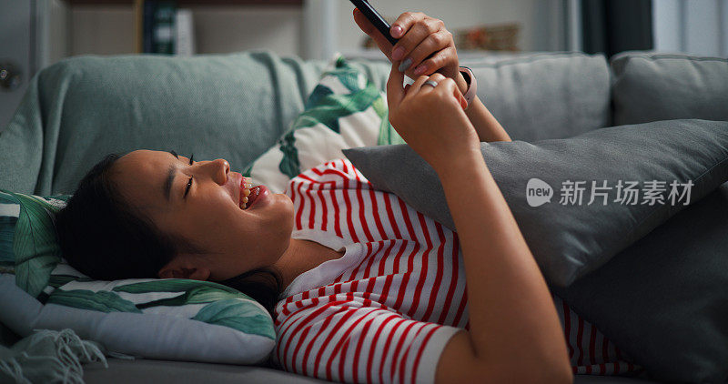 快乐的年轻女子一边笑一边在沙发上玩手机。