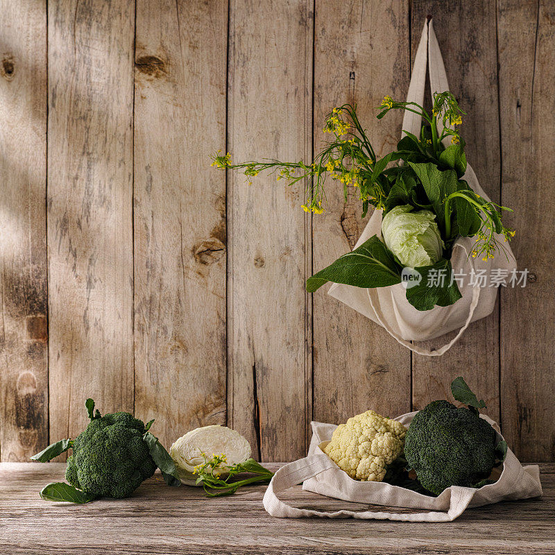 新鲜的绿叶蔬菜装在一个棉花可重复使用的购物袋里，用旧钉子挂在旧风化的木板墙上，还有更多的蔬菜放在袋子下面的木桌上。