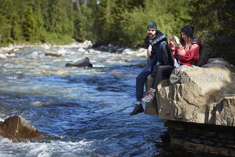 一对夫妇在河上的岩石上休息。女人用智能手机拍照