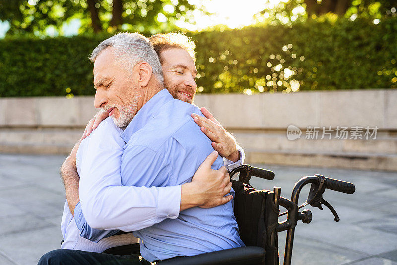 那位坐在公园里轮椅上的老人身边来了他的儿子。他正在拥抱那个老人
