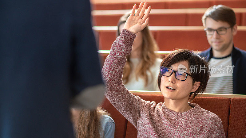在课堂上，聪明的亚洲学生举手向讲师提问。多元种族的现代聪明学生在学院。