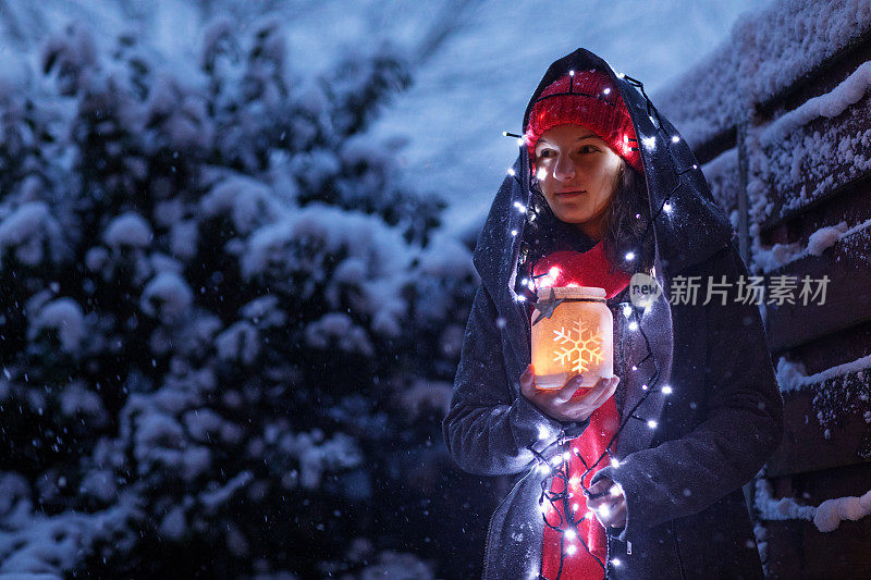 美丽的黑发少女穿着连帽大衣，同时在一个下雪的冬天晚上在户外举着圣诞灯