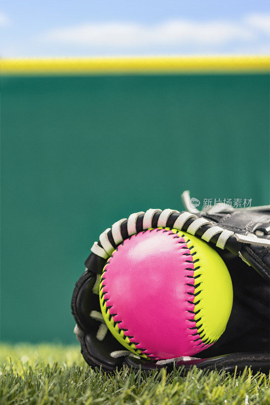一个黄色和粉红色垒球在一个黑色和白色垒球手套坐在合成草皮与墙的特写