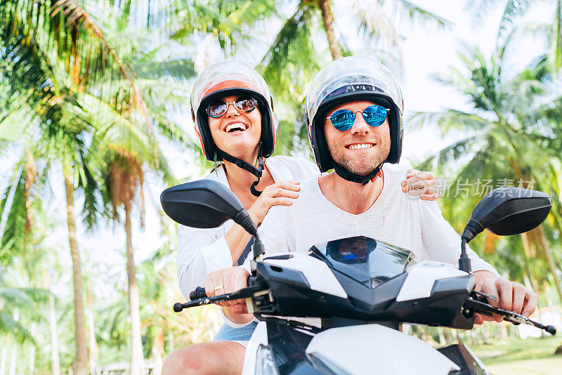 快乐微笑的夫妇旅行者骑摩托车在安全头盔在棕榈树下热带度假