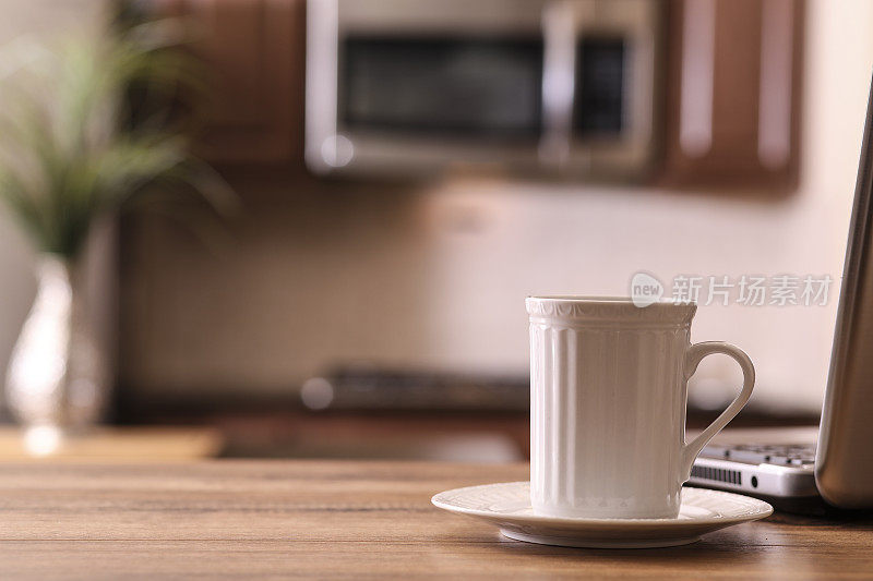 离焦厨房，咖啡杯，笔记本电脑在木头桌子的前景。