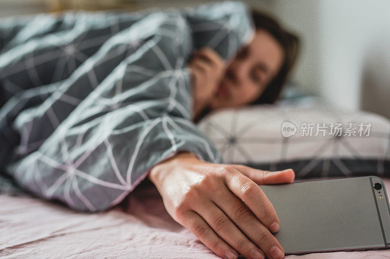 一个年轻的女人从床上醒来。智能手机上的闹钟响了