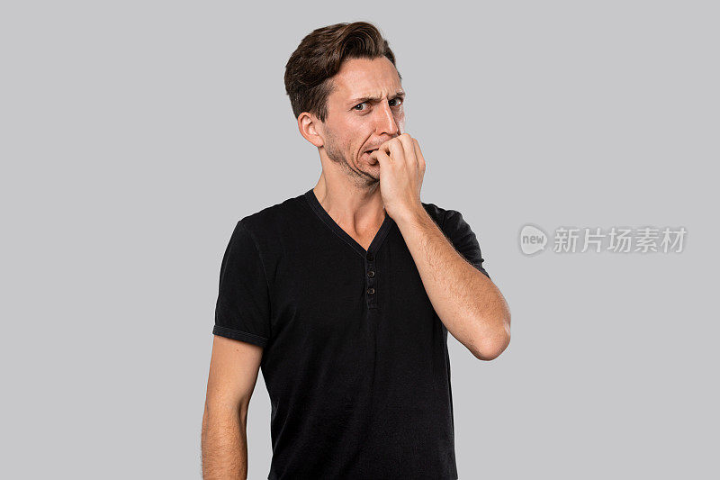 穿着黑色t恤的人咬他的指甲感到焦虑孤立在灰色的背景。