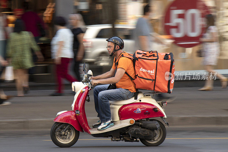 快递员骑着一辆红色和米色相间的老式小摩托车在城市的街道上行驶