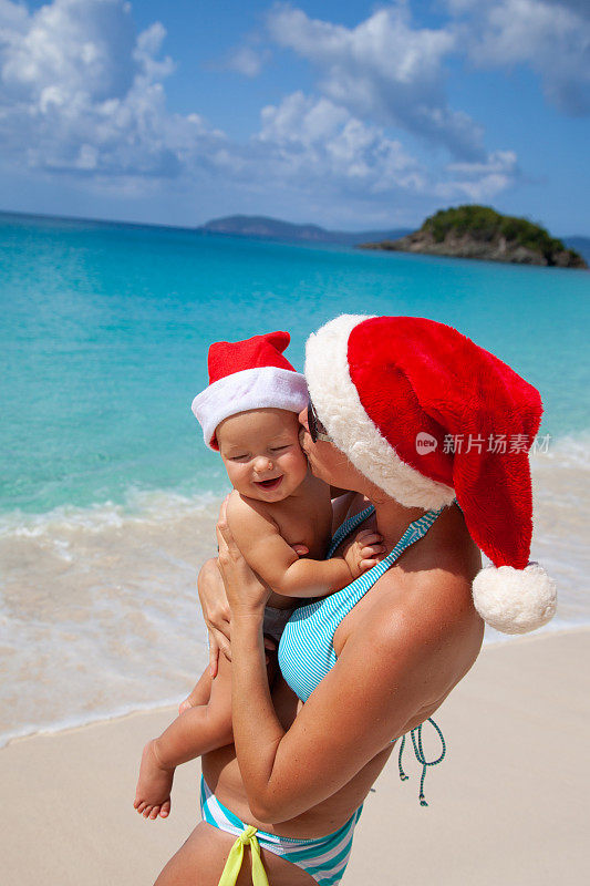 妈妈抱着快乐的婴儿戴着圣诞帽在加勒比热带海滩上