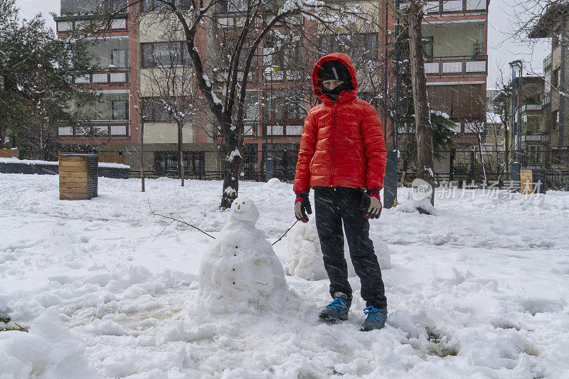 下雪天，穿红外套的男孩在操场上堆了一个雪人。