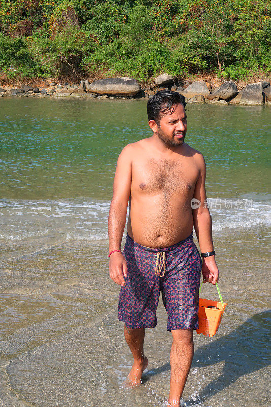 一名印度男子在海滩度假，背着塑料桶，从印度洋返回海滩，海浪翻滚到沙滩上，印度果阿巴洛伦海滩