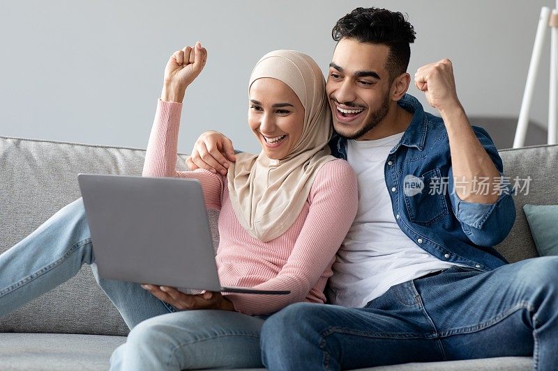 一对情绪激动的阿拉伯夫妇拿着笔记本电脑举起拳头庆祝成功