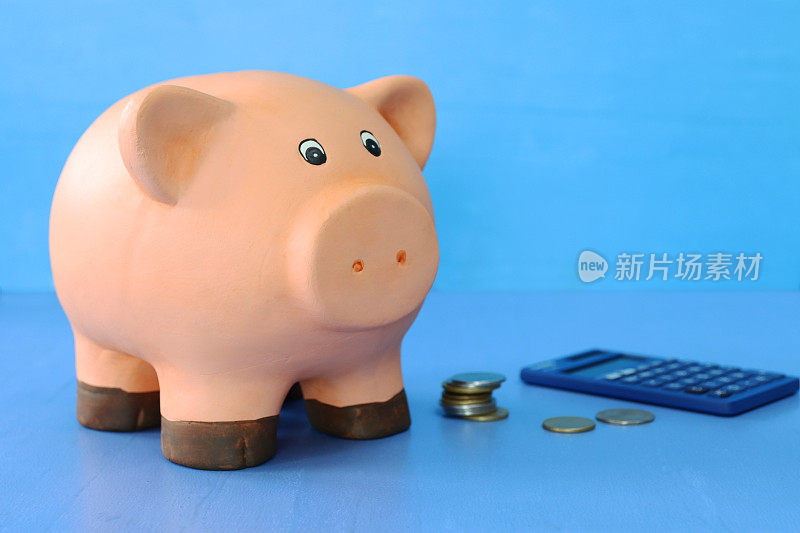 近景的小猪储蓄罐坐在计算器和成堆的硬币，蓝色背景，聚焦前景，抵押，家庭财务和储蓄的概念，复制空间