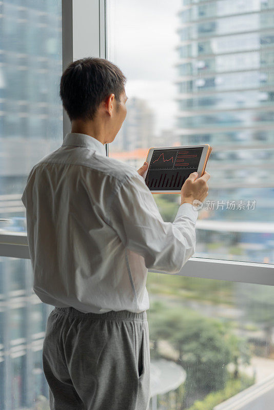 一个亚洲人站在窗前，用着平板电脑
